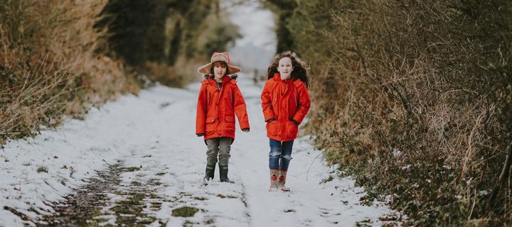 Two school-age children in bright orange coats walking along a snow-covered dirt road, dry brush on either side. 