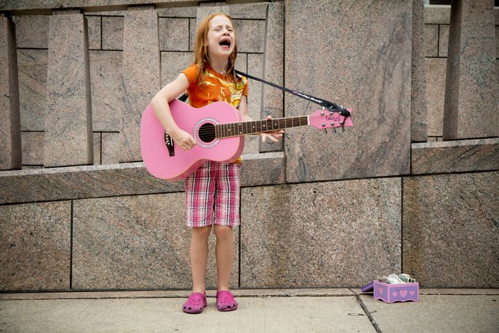 A child strums a pink guitar in front of a stone building, with a box to collect bills. 