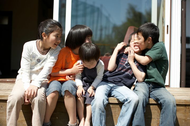 Five children seated on a deck. Two kids are fighting or mock-fighting, two watching and smiling, and the littlest, in the center, is ignoring the action.