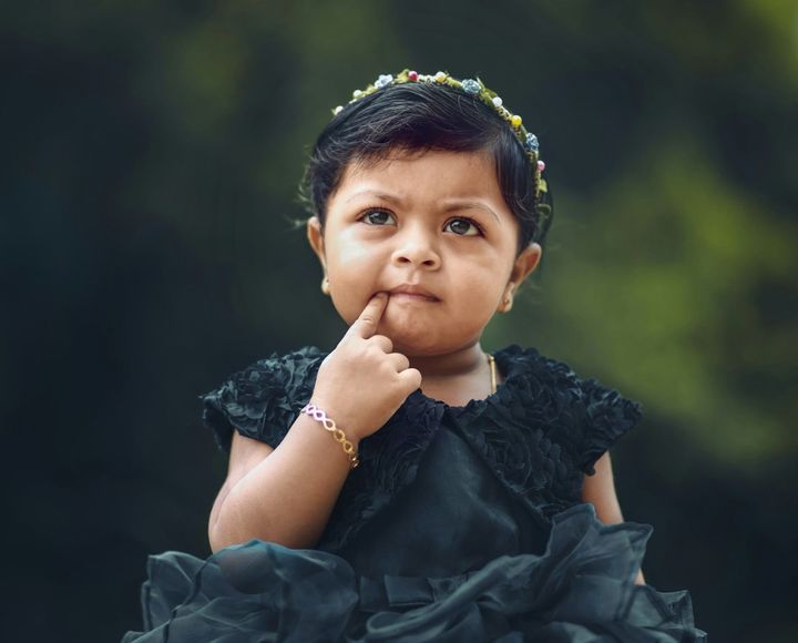 A toddler dressed in finery—flower headband, gold bracelet and earrings, elaborate black dress—has a finger to her mouth and a pondering expression.