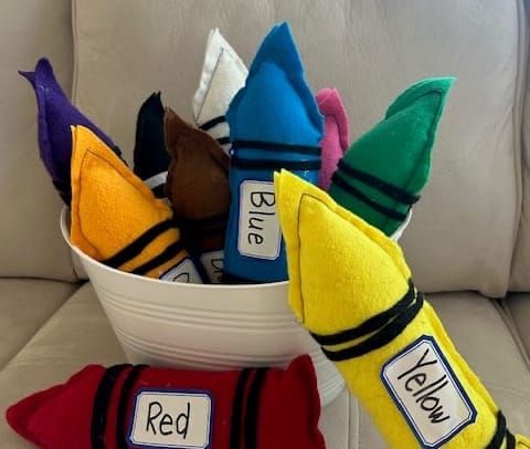 Stuffed felt crayon lookalikes in a bucket on a couch. Red and yellow "crayons" outside the bucket.