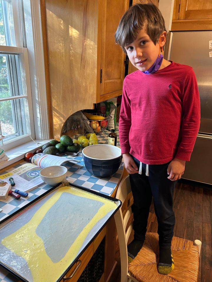 A boy stands on a chair in a kitchen. On the counter is a sheet pan filled with multicolored batter for a "tie-dyed roll cake." 