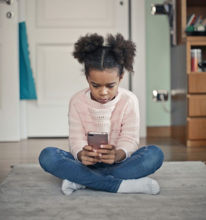 A child sits cross-legged alone indoors using a smartphone.