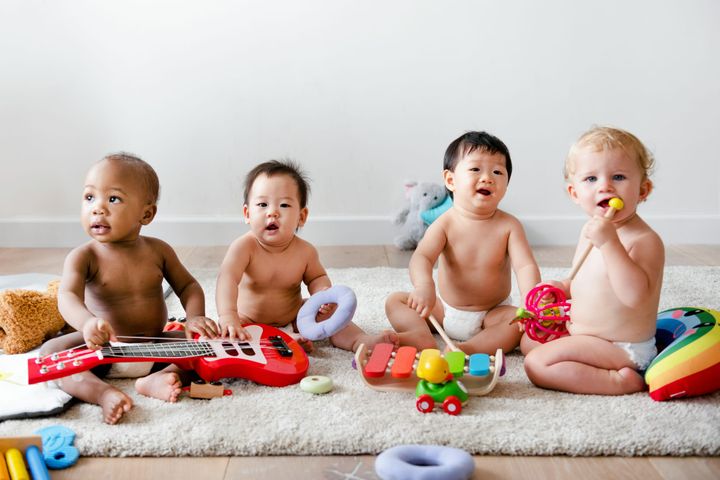 Four babies sit on a fuzzy rug with various toy musical instruments.