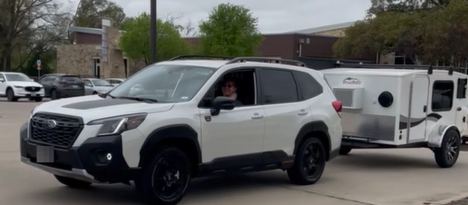 White Subaru SUV towing a small white camper trailer, with driver visible through window, parked in a parking lot.