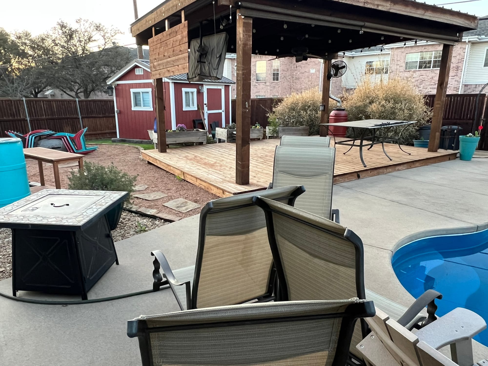 Backyard wooden deck with pergola, outdoor furniture, and a small red shed surrounded by planters and a wooden privacy fence.