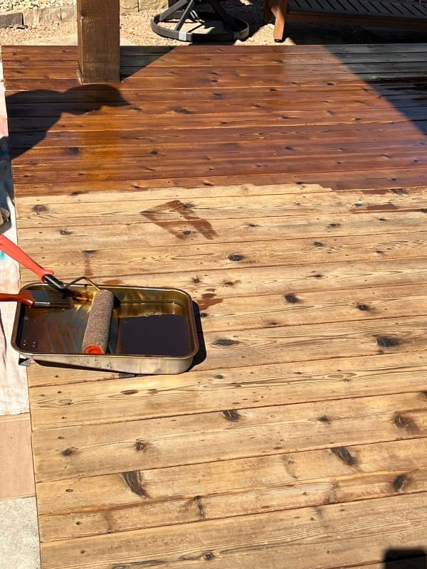 Wooden deck showing transformation in progress with half stained in rich brown finish and half unstained, with staining roller and tray visible in the foreground.