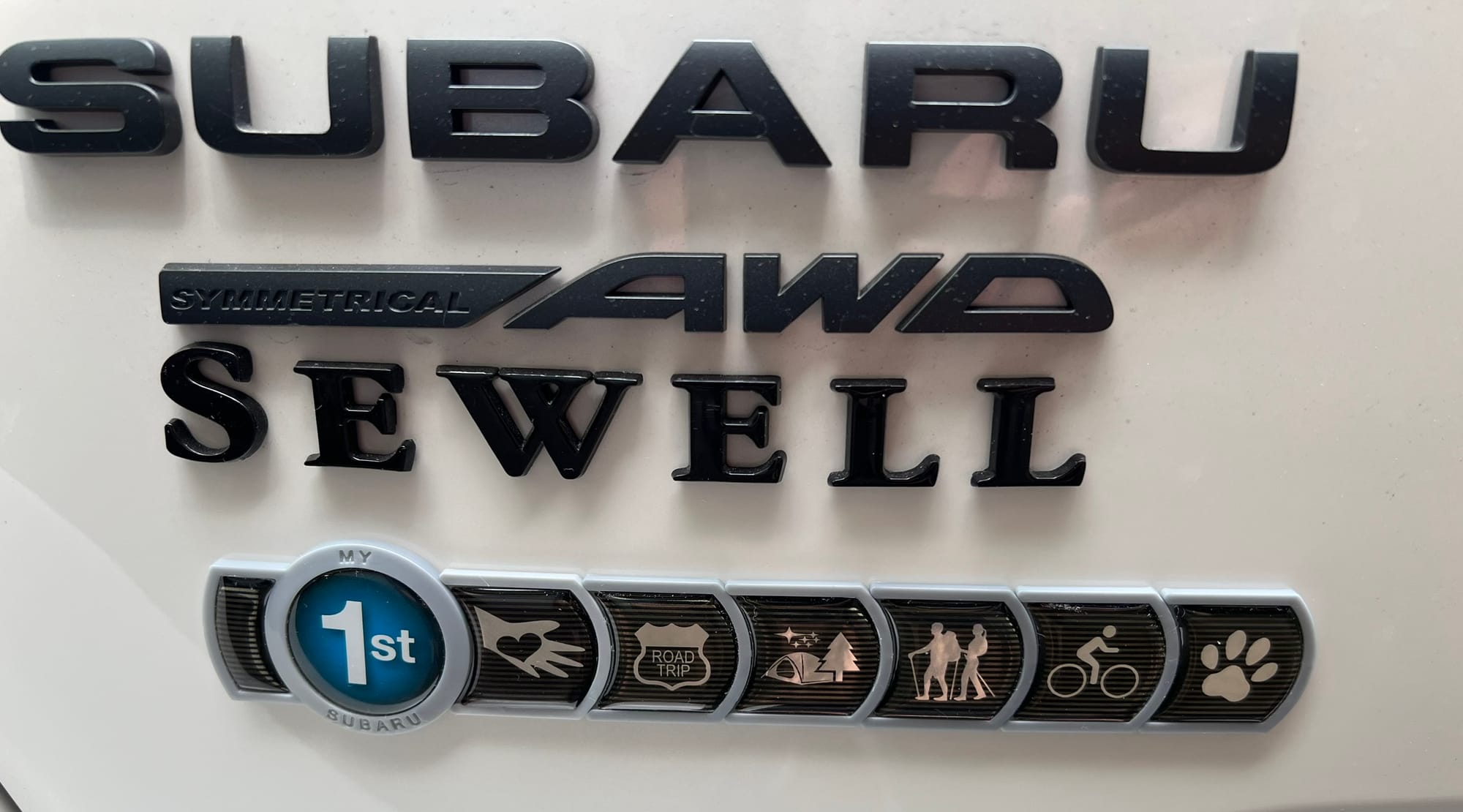 Close-up of Subaru emblem on white car with "1st" badge and lifestyle badges showing road trip, camping, hiking, cycling, and pet ownership icons.