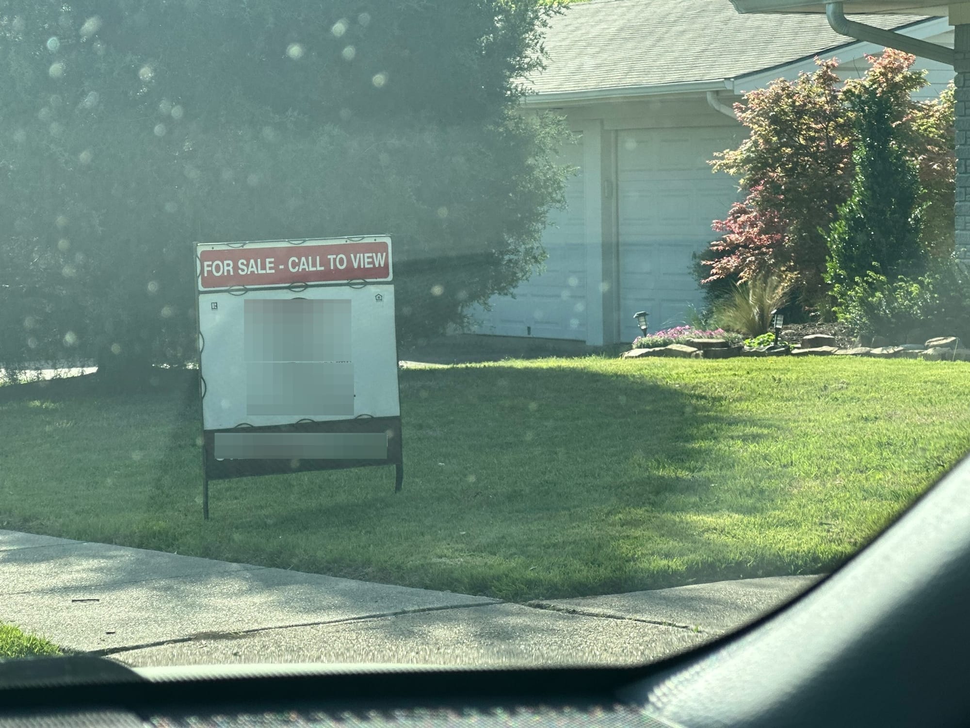 A "For Sale" sign in a well-maintained front yard with green grass, viewed through a car window.