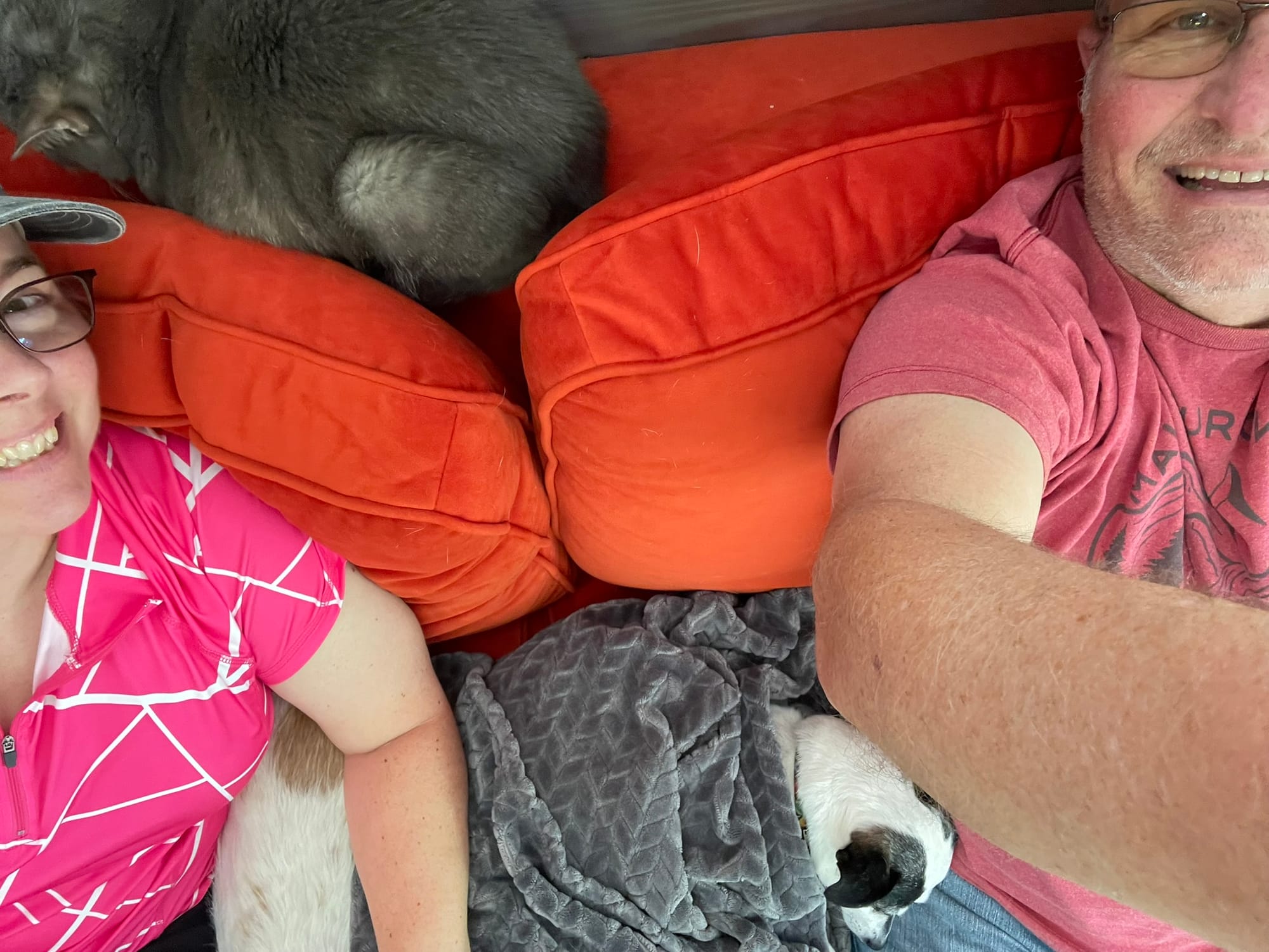 A cozy scene showing a cat and dog snuggled together with humans on a small couch in the "tinytel", with orange cushions and gray blankets.