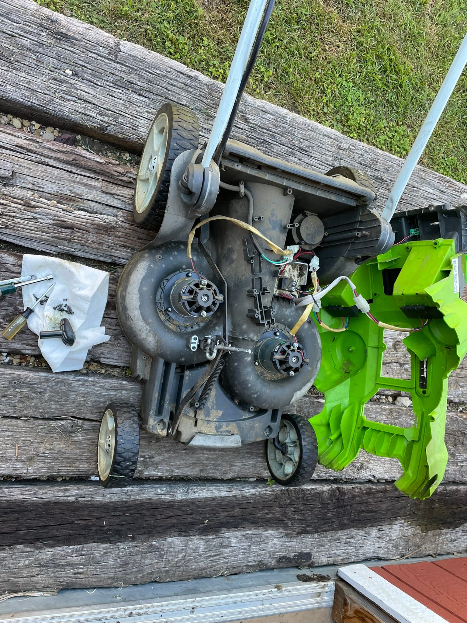 A lawn mower showing the mechanical components during repair work. Tools are scattered nearby on wooden decking.