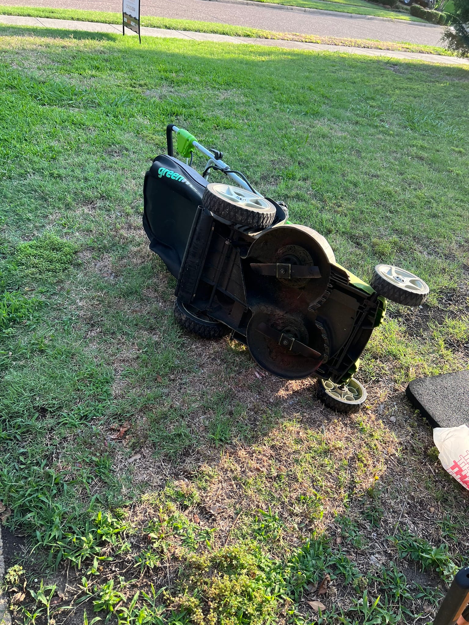 Green and black push mower tipped over on grass, appearing broken and abandoned
