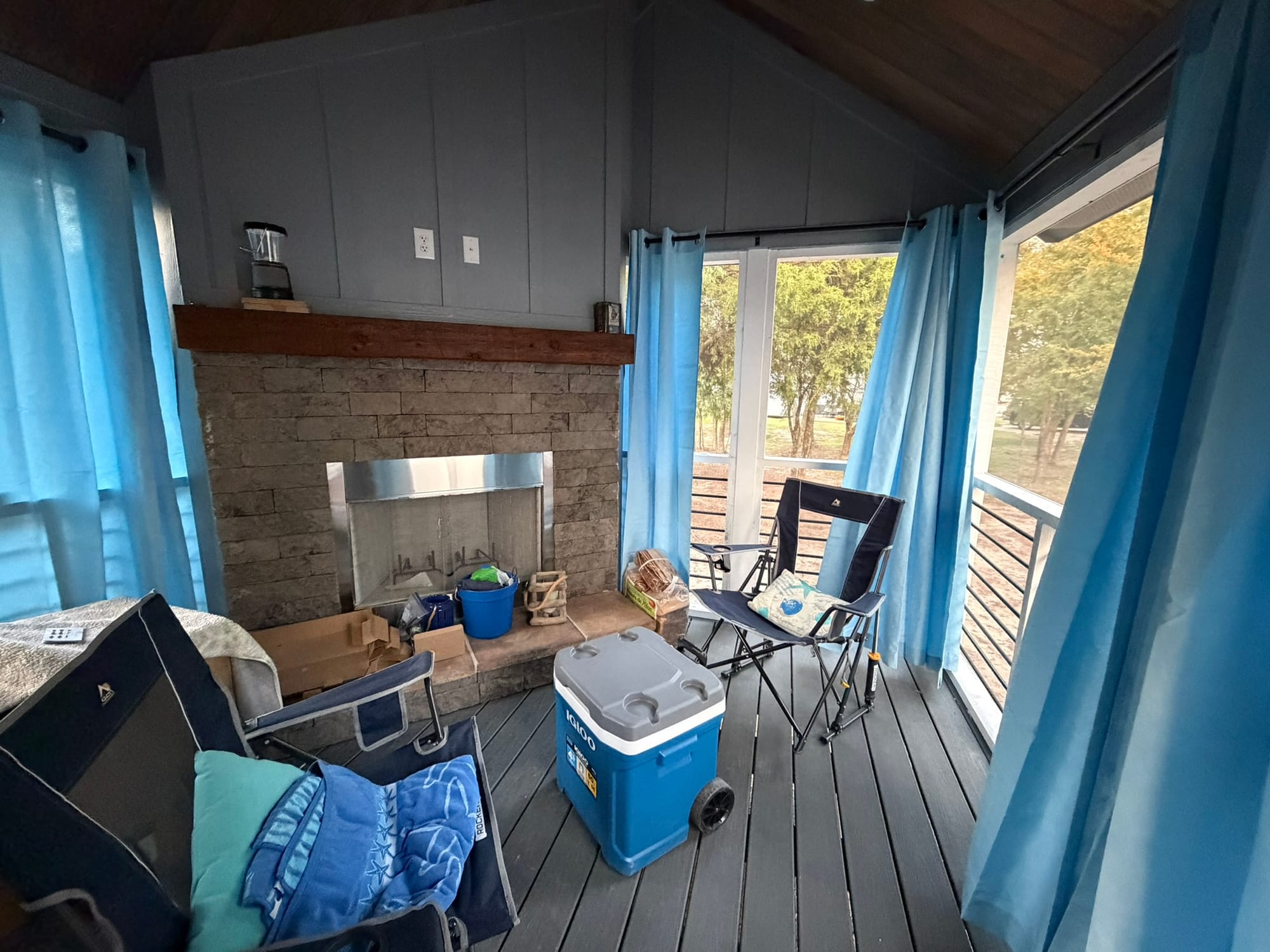 Covered porch with blue curtains hung for privacy, camping chairs and cooler visible, showing functional outdoor living space