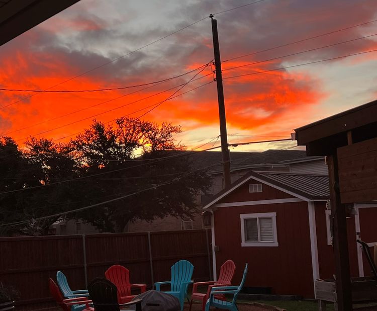 Vibrant orange and pink sunset sky over backyard with colorful Adirondack chairs and small shed, creating a peaceful outdoor living space.