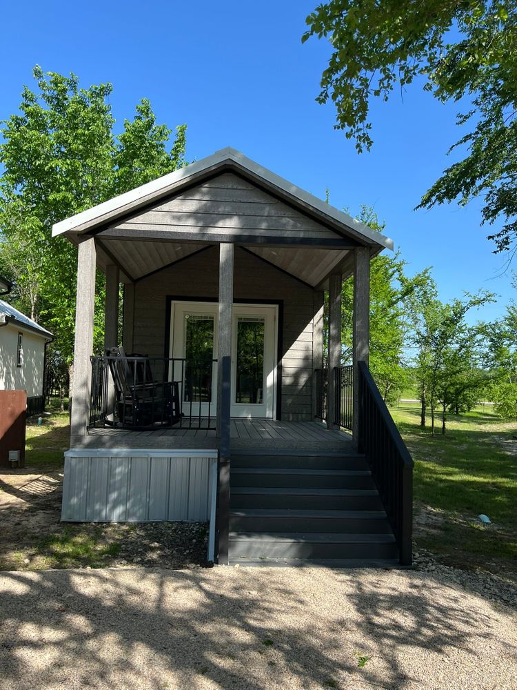 Exterior view of a small gray tiny home with covered porch, white door, windows, and stairs leading to the entrance, surrounded by trees.