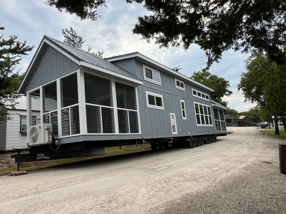 Gray tiny house with white trim and screened porches sitting on a community road, positioned at an angle with trees in the background