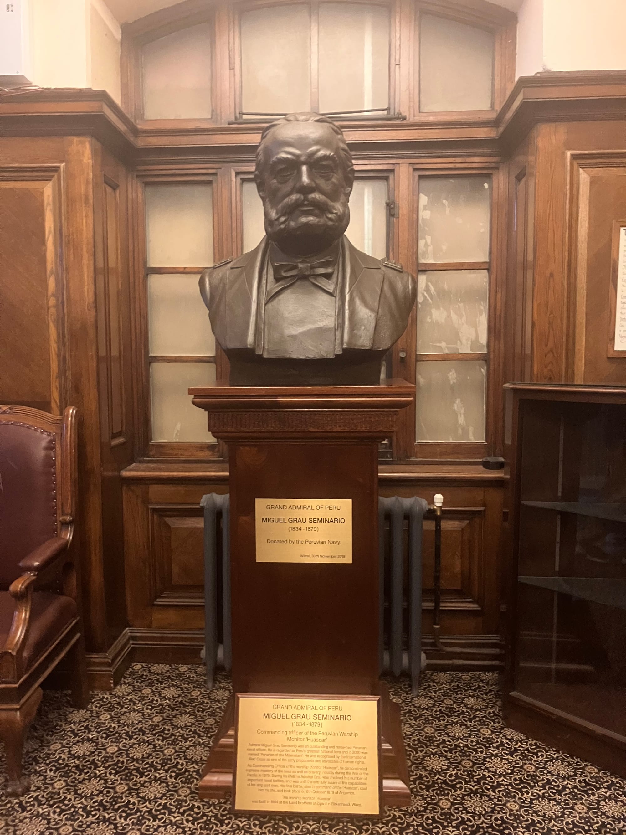 Image of a bust of Miguel Seminario located in Wallasey town hall. He was the grand admiral of Peru. The bust is of bronze appearance nd sits on a wooden plinth. Below a patterned carpet, to the left a wood and leather lounge chair and behind is wood panelling and glazing
