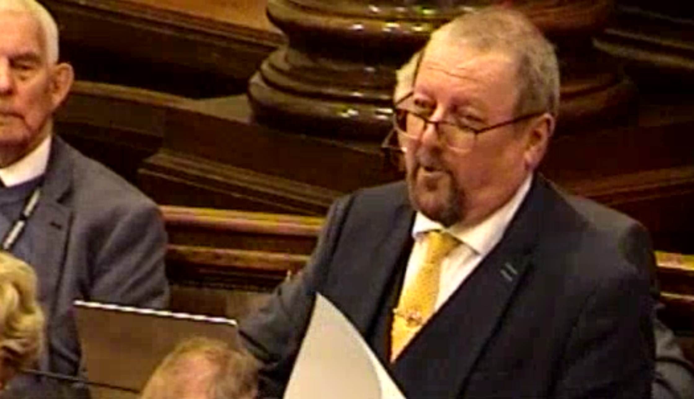 Screenshot Image from Wirral Council video of Cllr Carubia speaking at the meeting. He is smiling, standing, mid speech, paper is visible in his hands. Behind him the tops of heads of two councillors are visible, alongside a Reform councillor, who is seated