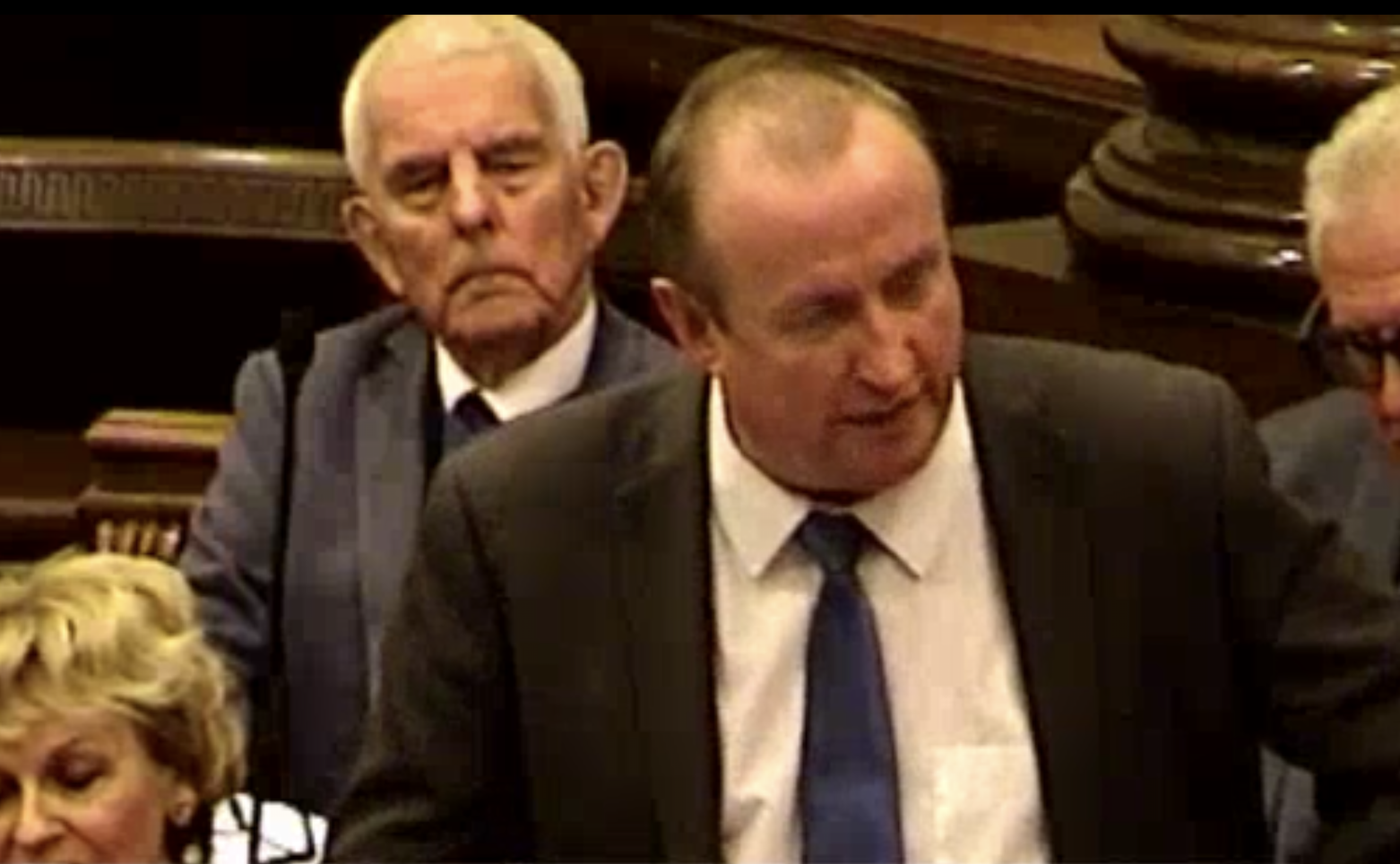 Screenshot Image from Wirral Council video of Cllr Kelly standing and speaking at the meeting. Behind him, seated are two Reform councillors