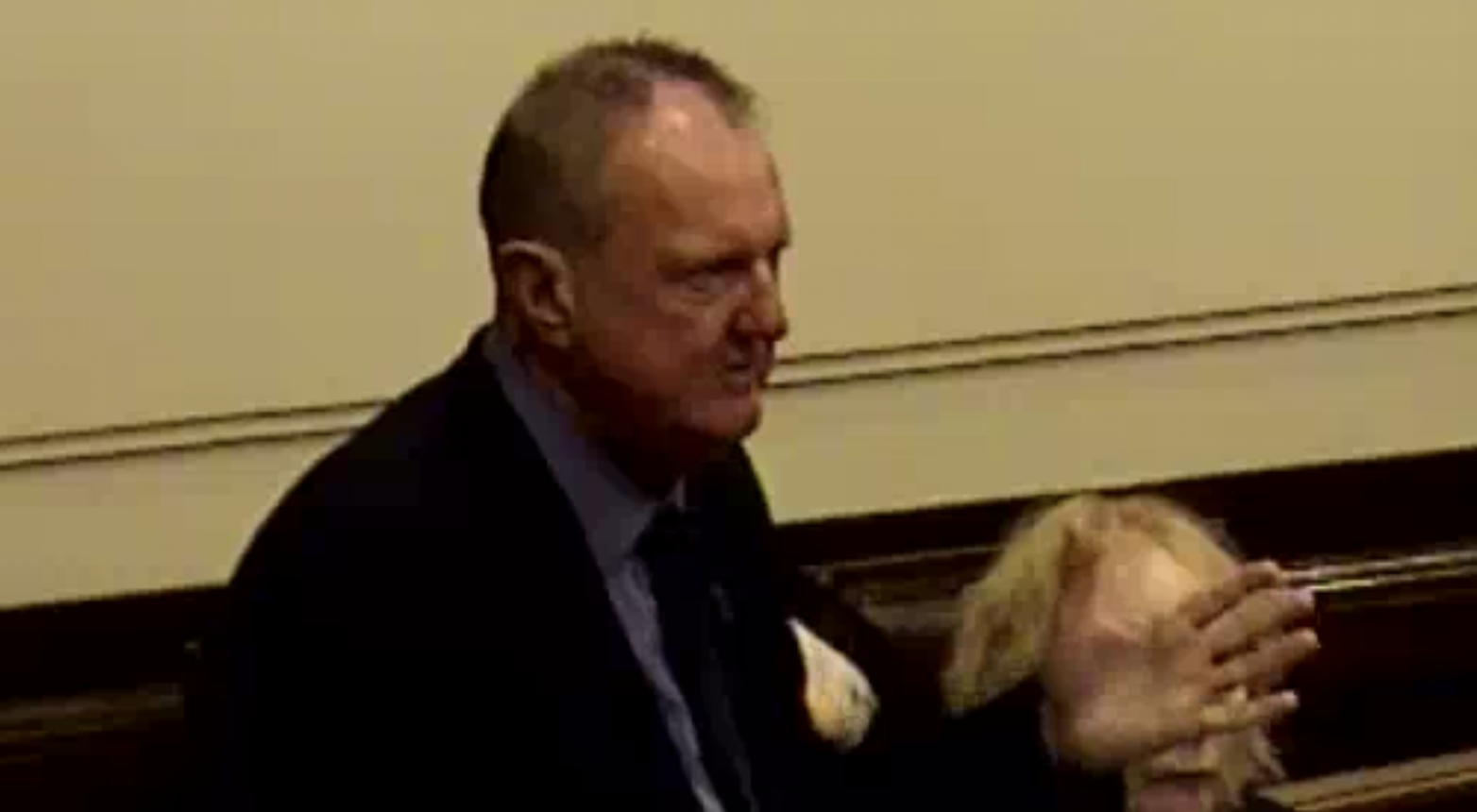 Screenshot image from Wirral Council video of Cllr Foulkes speaking at the meeting. His hand is outstretched and he is standing, behind him the obscured face of a fellow Labour councillors seated, who is seated.