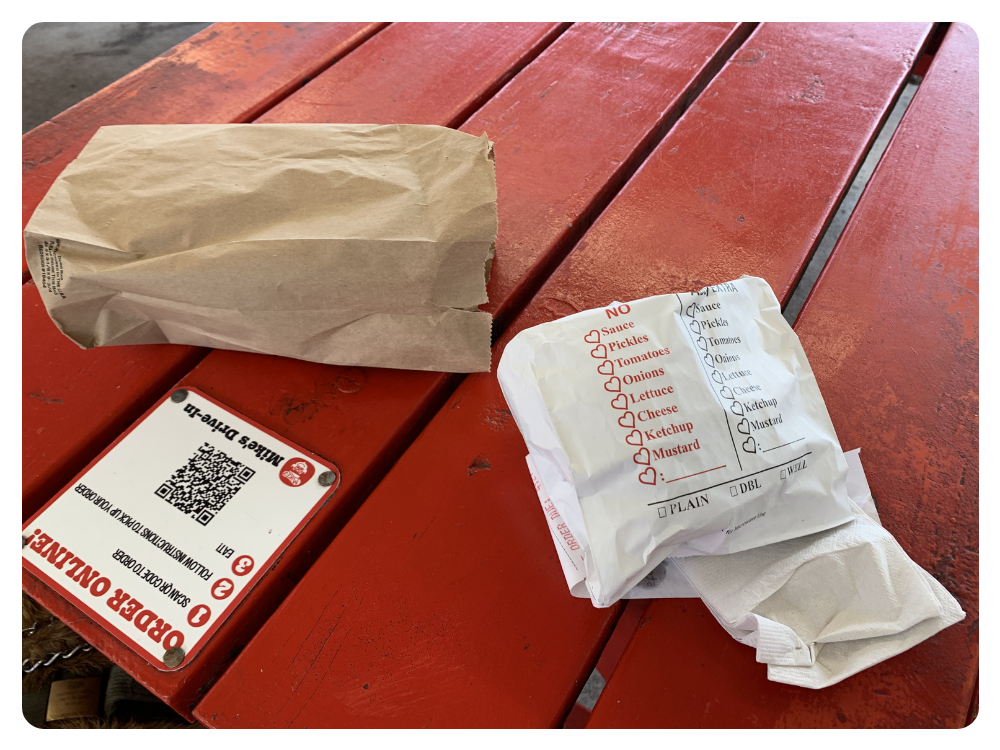 Close up of a red painted picnic table. A Mike's Drive In placard is screwed to the table encouraging "ORDER ONLINE" On the table is a brown paper sack. Directly next to it is a white paper pouch with the order printed on the side and some napkins.