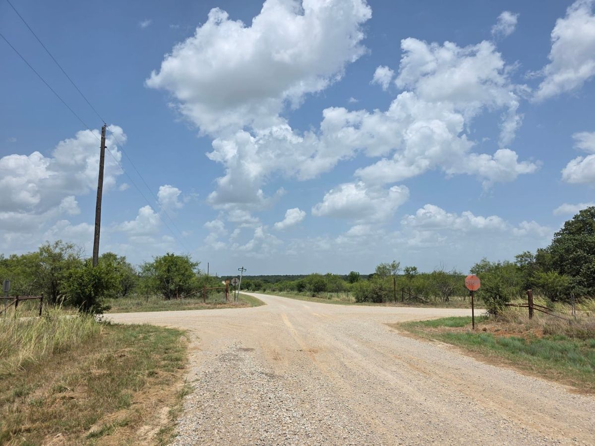 Gravel county roads meet at a rural Texas crossroads bordered by fence posts, brush and a faded stop sign beneath a wide blue sky.
