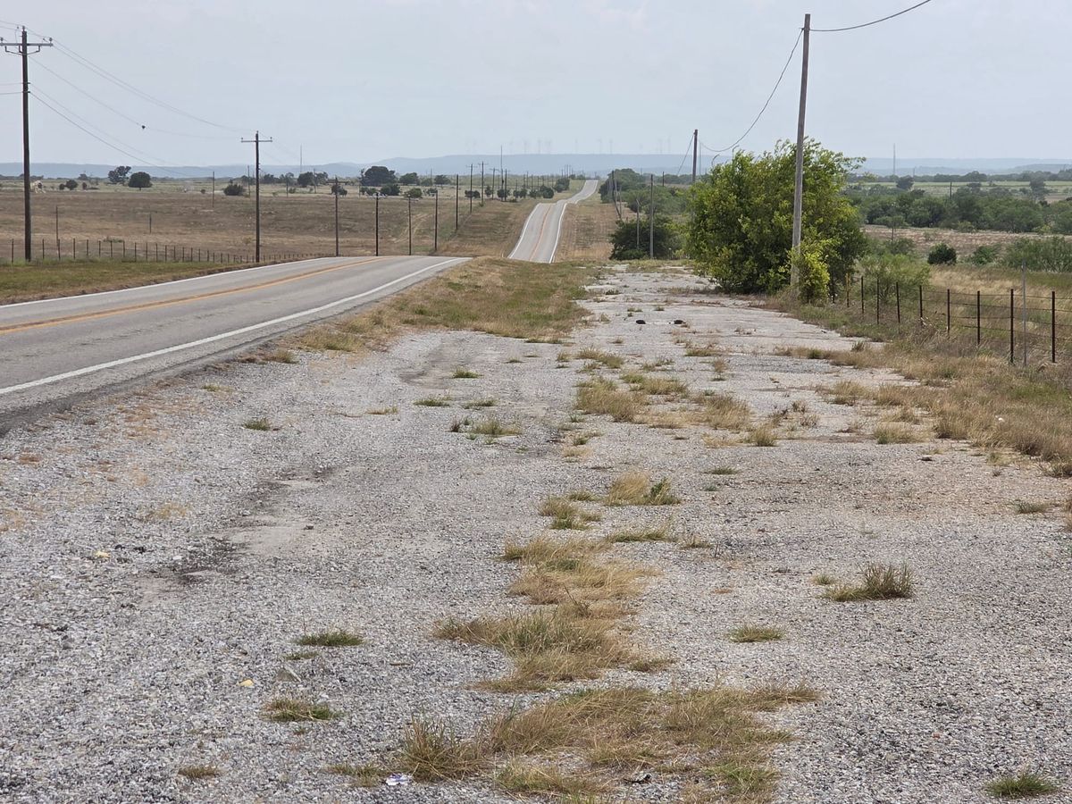 A long rural highway crosses open Jack County land beneath utility lines, with wind turbines visible along the distant ridge.