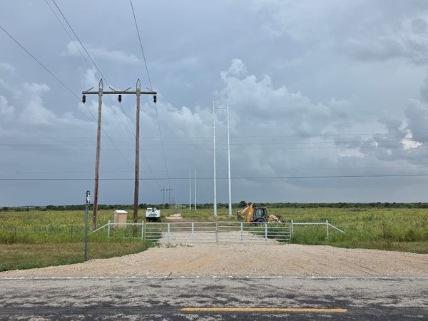 A rural Texas road and pasture beneath a wide sky with electrical transmission infrastructure crossing the landscape.