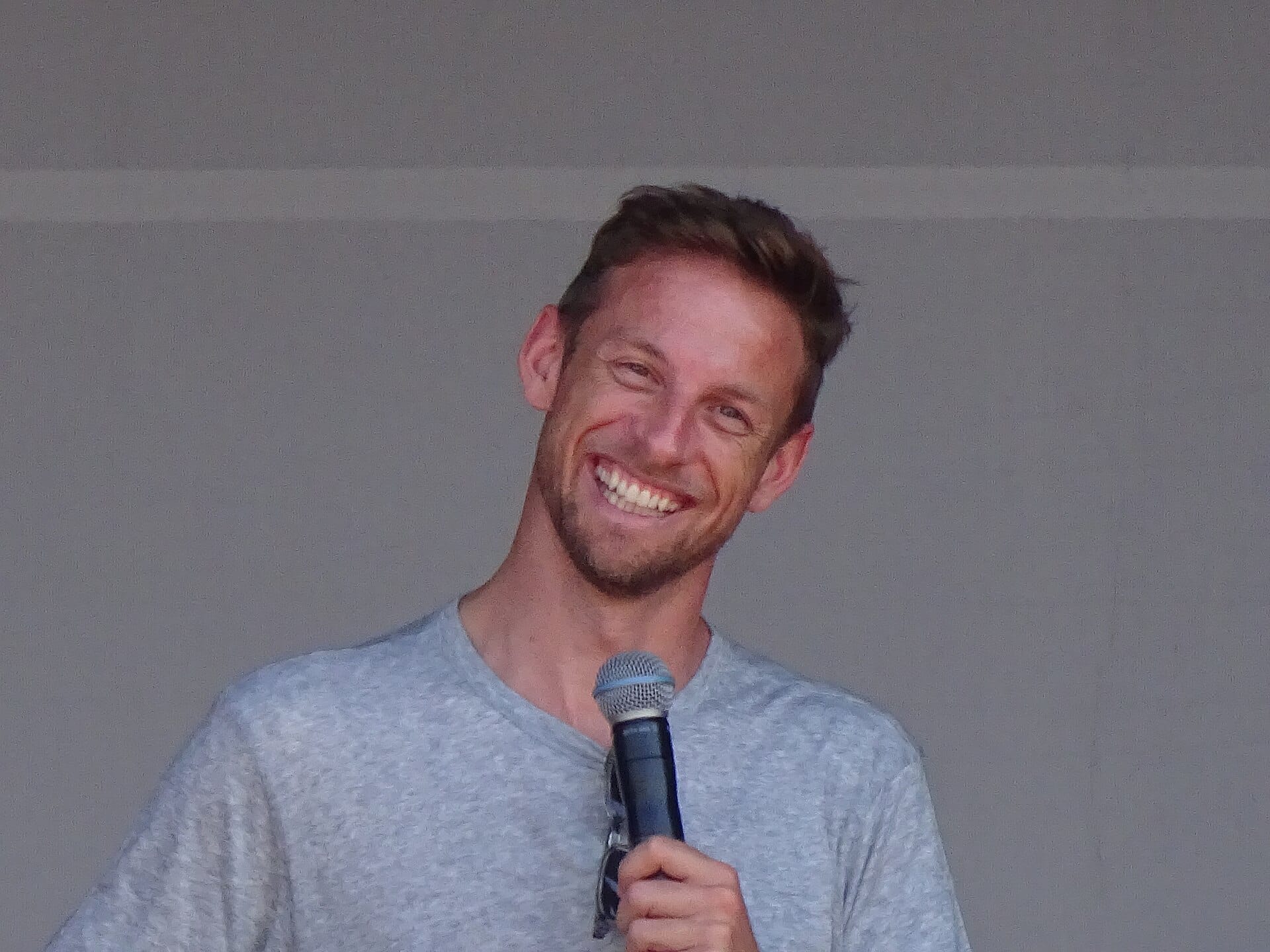A portrait of Jenson Button smiling cheerfully while holding a microphone.