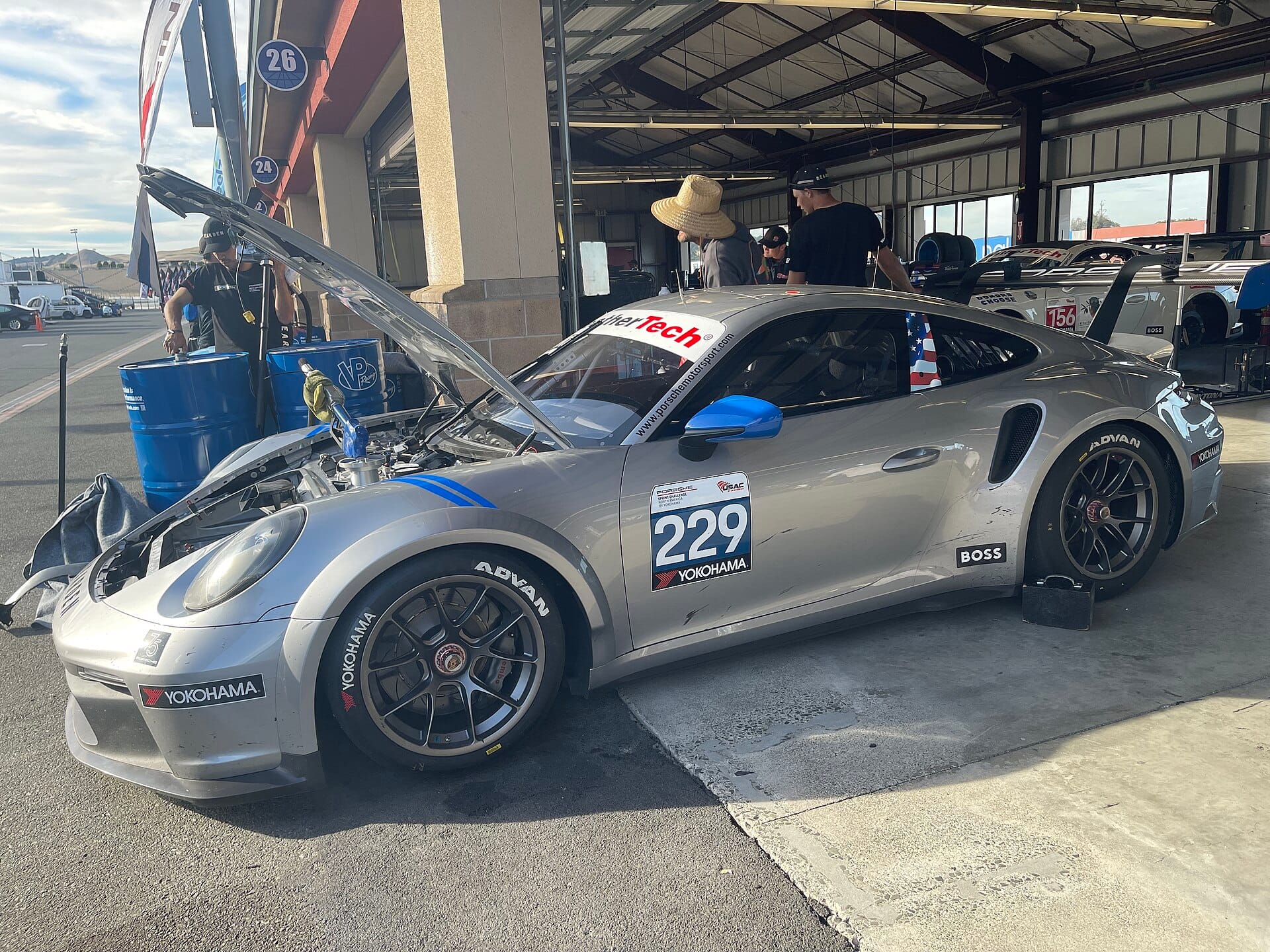 A silver Porsche 911 GT3 Cup race car with its hood open, parked in a pit garage area during a race event.