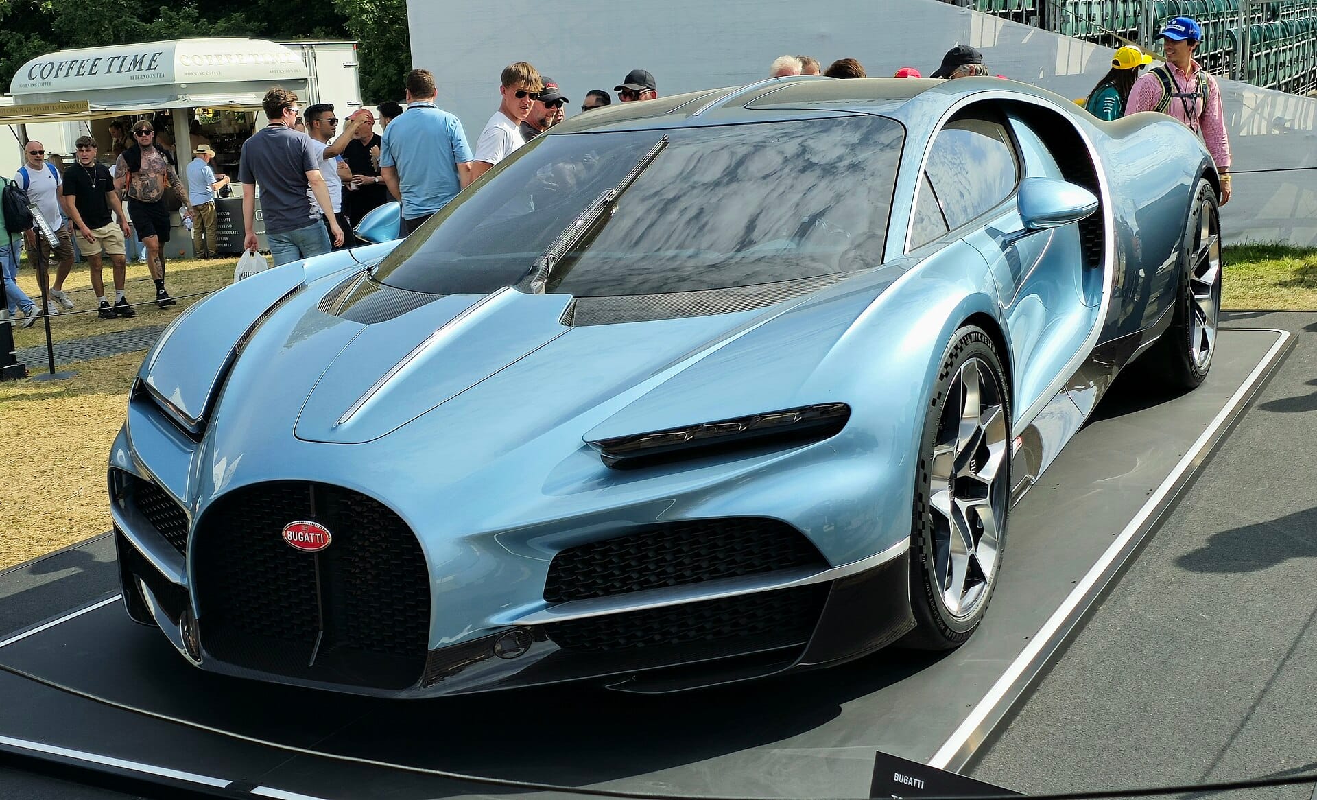 A front three-quarter view of a metallic light blue Bugatti Tourbillon hypercar on display outdoors with spectators in the background.