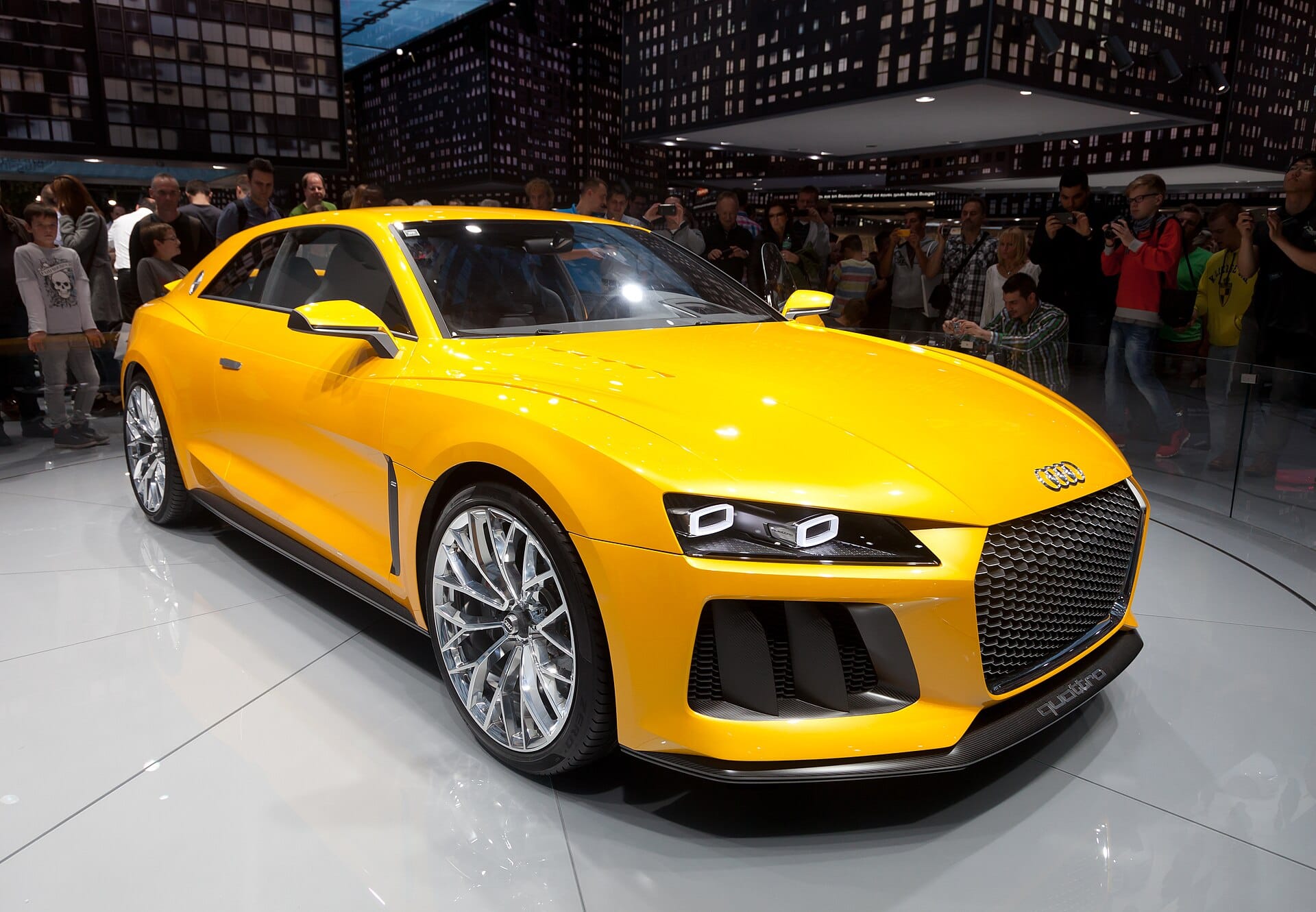A bright yellow Audi Sport quattro concept car displayed on a polished show floor at an auto exhibition surrounded by onlookers.
