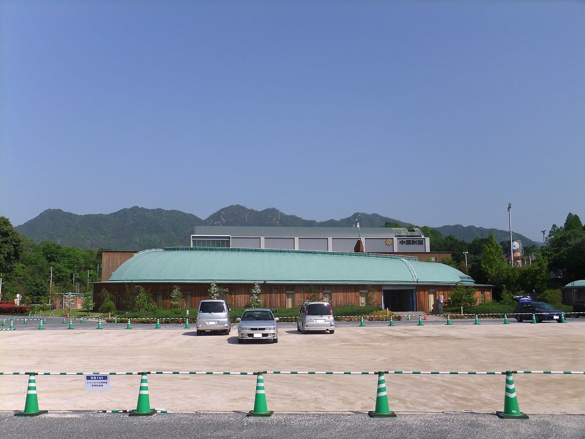 An exterior wide shot of a Chugoku Shimbun building with a green curved roof set against a backdrop of mountains under a clear blue sky.