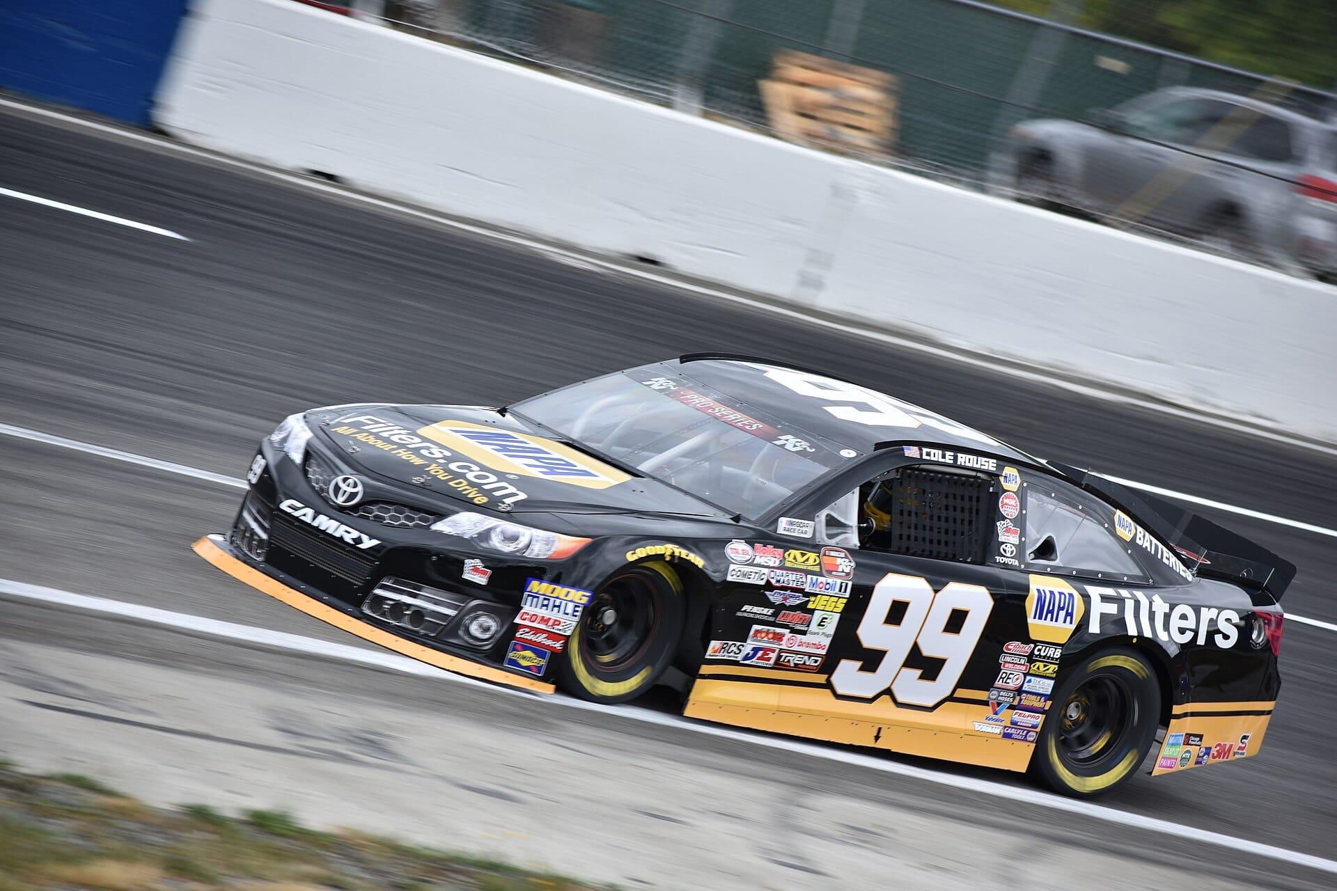 A black and yellow #99 Toyota Camry stock car racing around a paved oval track during a NASCAR Pro Series event.