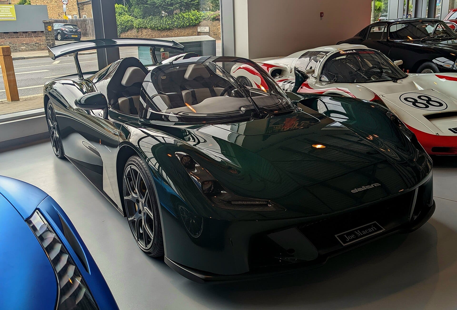 A dark green Dallara Stradale sports car on display inside a showroom alongside other high-performance vehicles.
