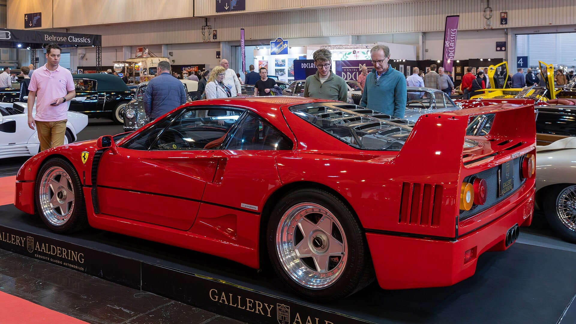 A bright red Ferrari F40 on display at a classic car show by Gallery Aaldering, with spectators admiring it in the background.
