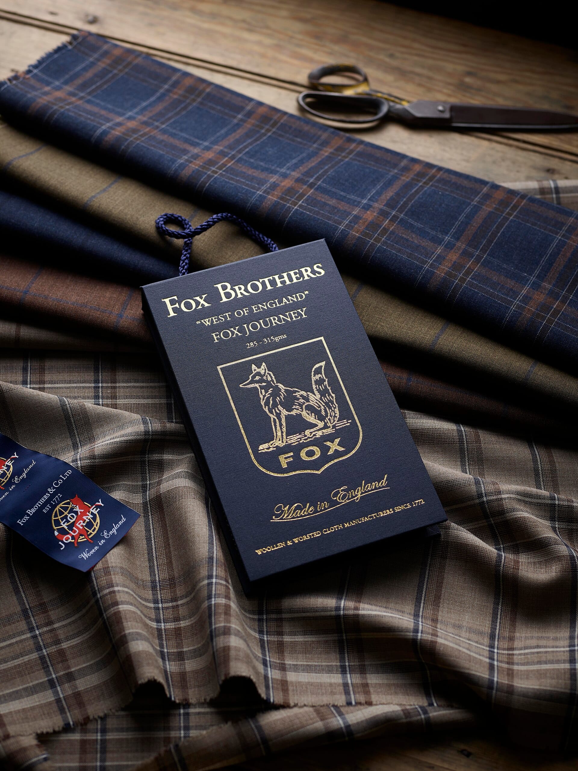 A high-angle product photograph featuring a dark blue Fox Brothers fabric bunch book surrounded by woolen plaid swatches and tailoring scissors.