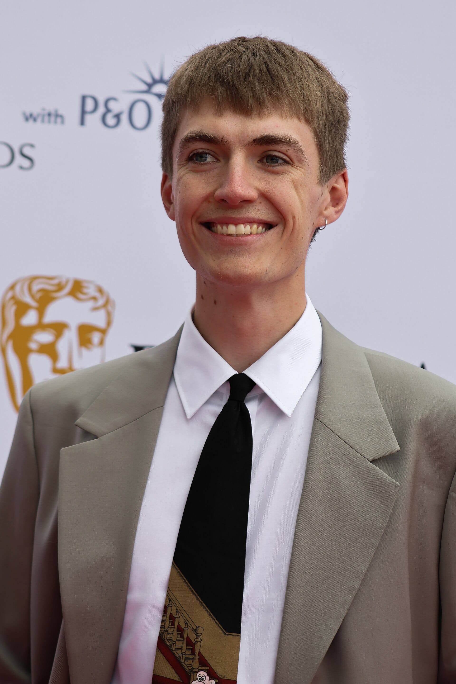 A close-up portrait of Francis Bourgeois wearing a grey suit jacket, white shirt, and black tie smiling at a red carpet event.
