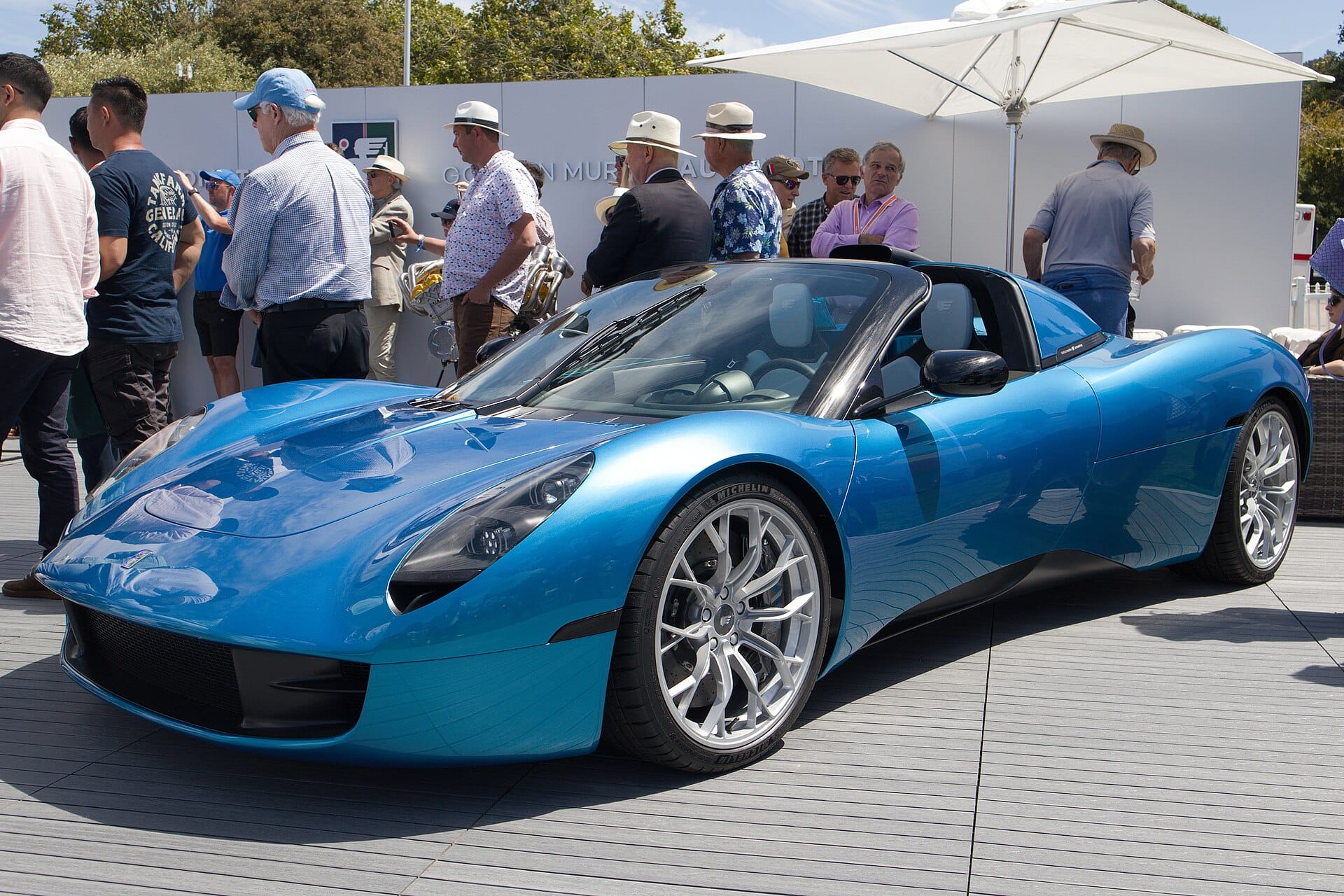 A metallic blue Gordon Murray Automotive T.33 Spider displayed outdoors on a deck at an automotive event surrounded by spectators.