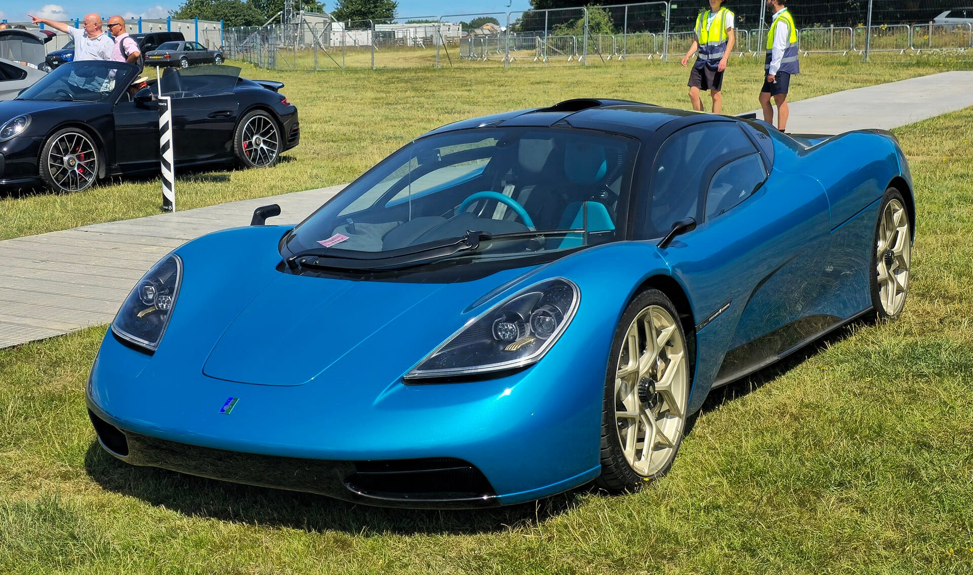 A front three-quarter view of a metallic blue Gordon Murray Automotive T.50 supercar parked on grass at an outdoor automotive event.