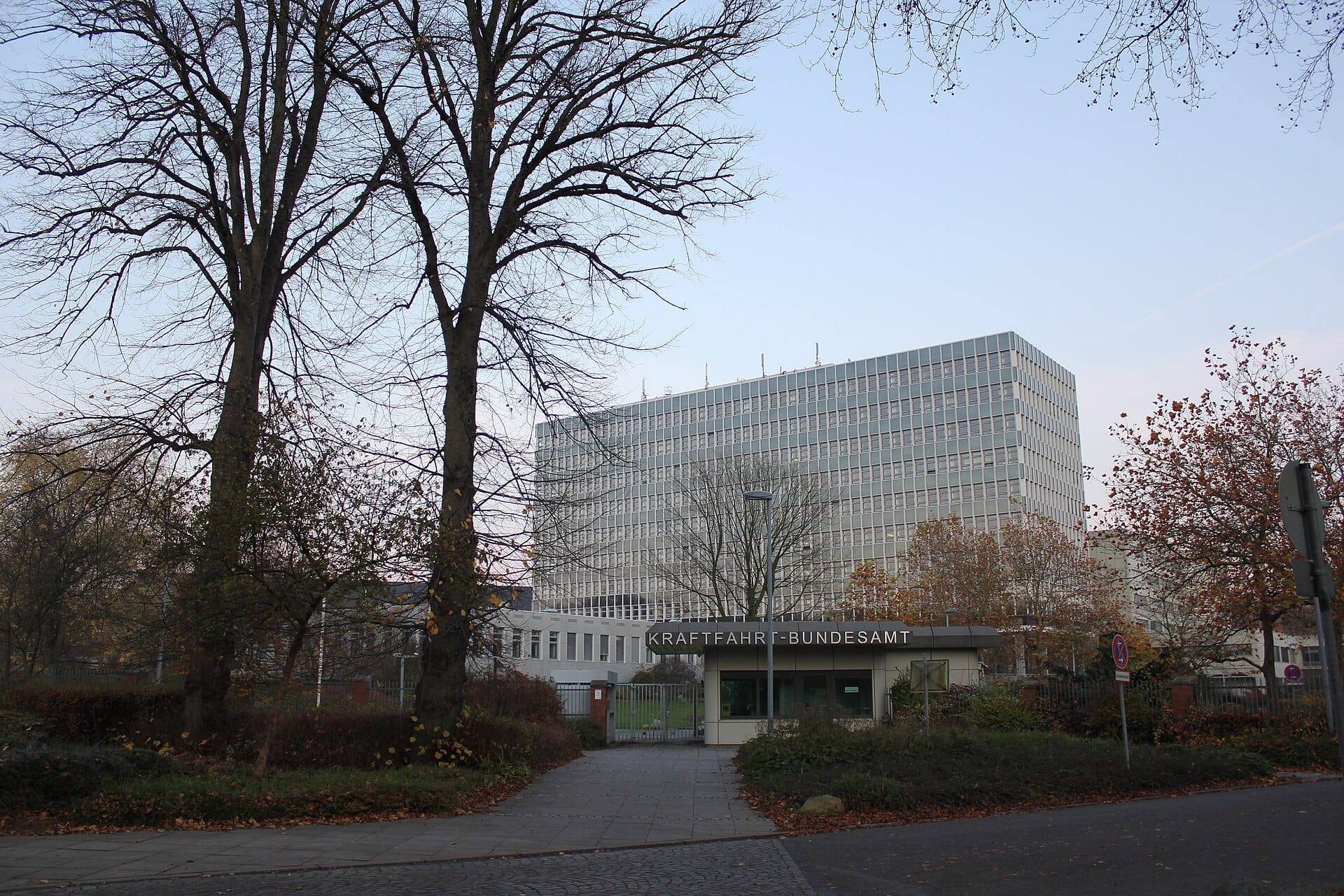 An exterior view of the Kraftfahrt-Bundesamt headquarters building in Flensburg showing the entrance gatehouse with visible signage and autumn foliage.