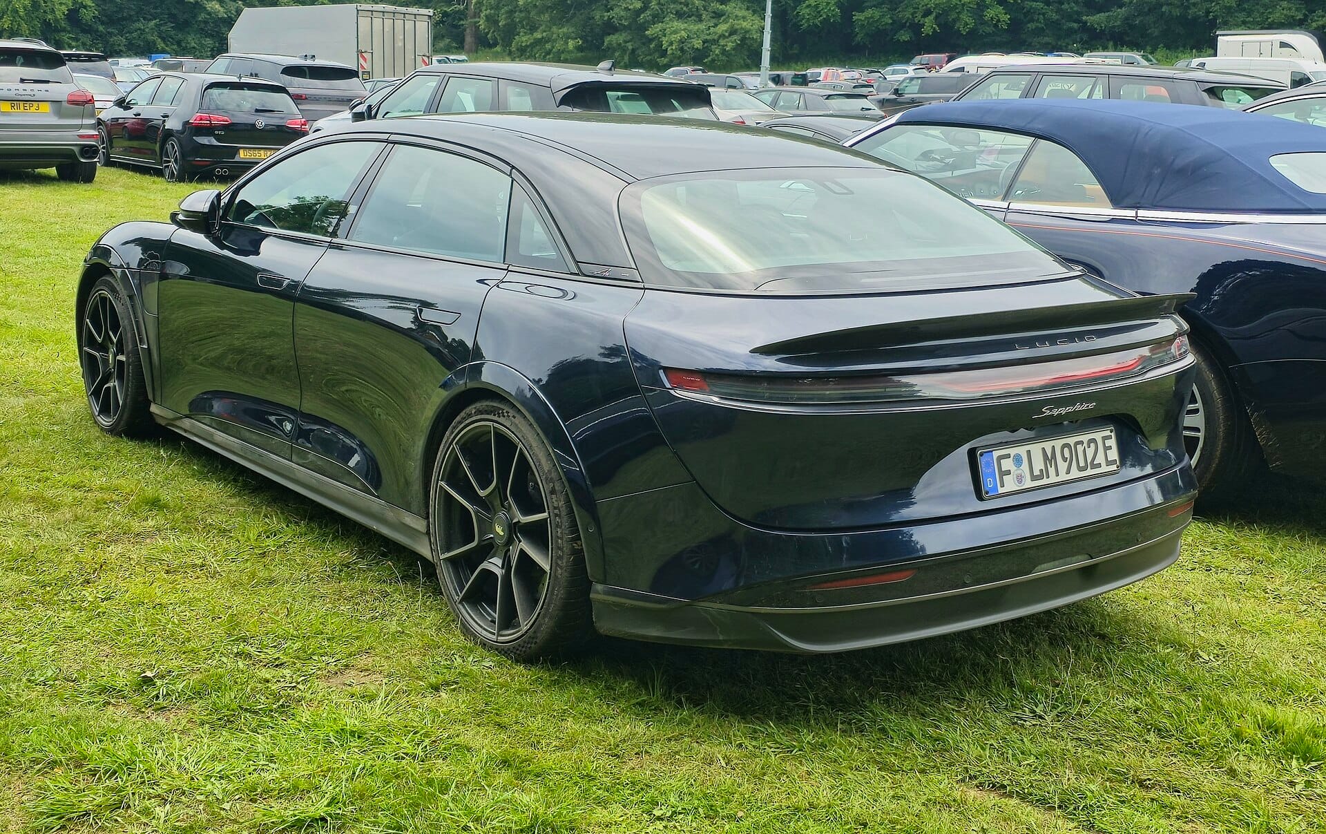 A rear three-quarter shot of a dark blue Lucid Air Sapphire parked on a grassy field alongside other cars at an outdoor event.
