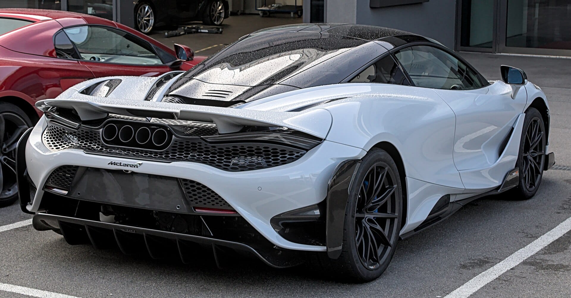 A rear three-quarter view of a light gray McLaren 765LT supercar parked in a lot, featuring four central exhaust pipes and aggressive aerodynamic bodywork.