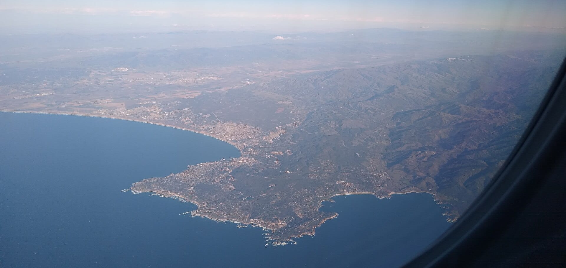 An aerial photograph taken from an airplane window showing the Monterey Peninsula, Carmel Bay, and the surrounding California coastline.