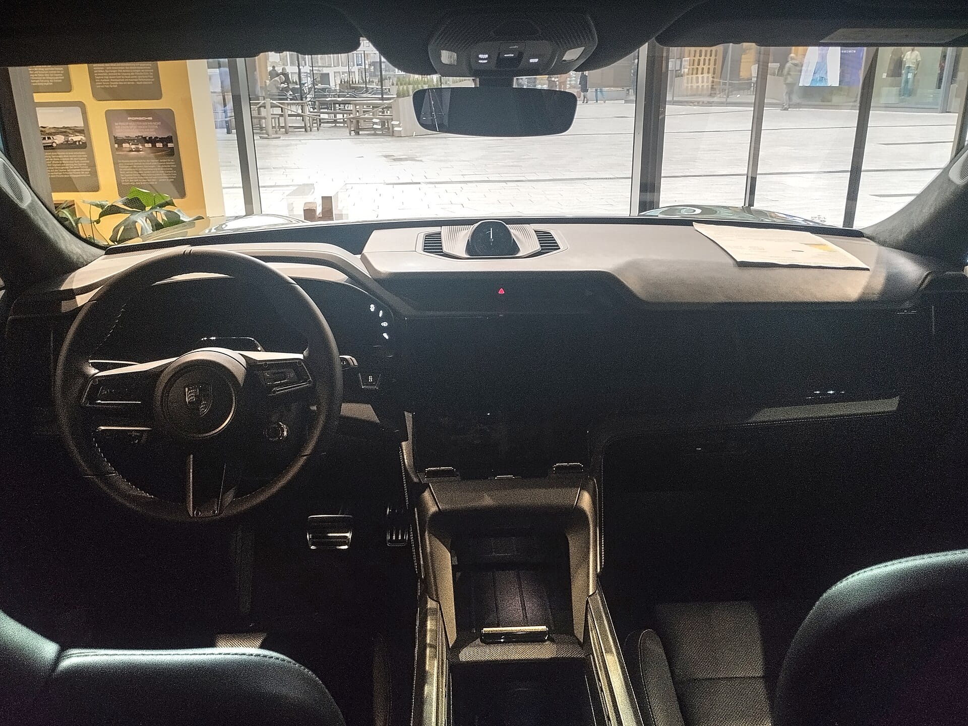An interior shot from inside a Porsche vehicle, displaying the dark dashboard, steering wheel with the Porsche crest, digital instrument cluster, and central console looking out through the front windshield.