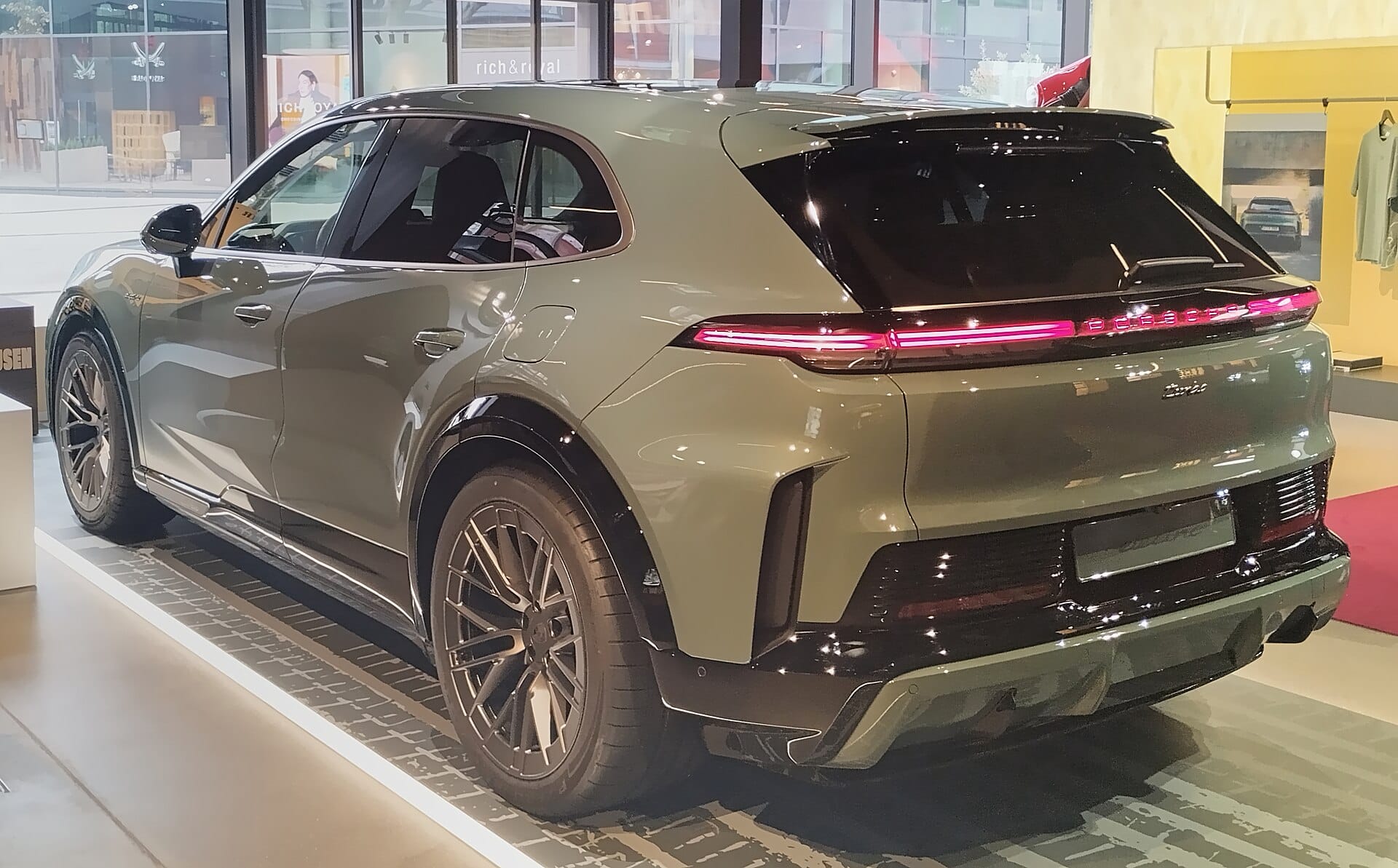 A rear three-quarter view of an olive green Porsche SUV displayed inside a showroom featuring a light bar across the tailgate.