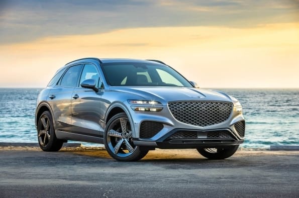 A silver Genesis GV70 luxury SUV parked near the ocean shore during sunset, captured from a front three-quarter angle.