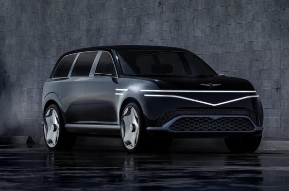 A dark grey Genesis concept SUV parked against a textured grey wall, featuring two-line front LED headlights that form the brand's iconic crest grille shape.