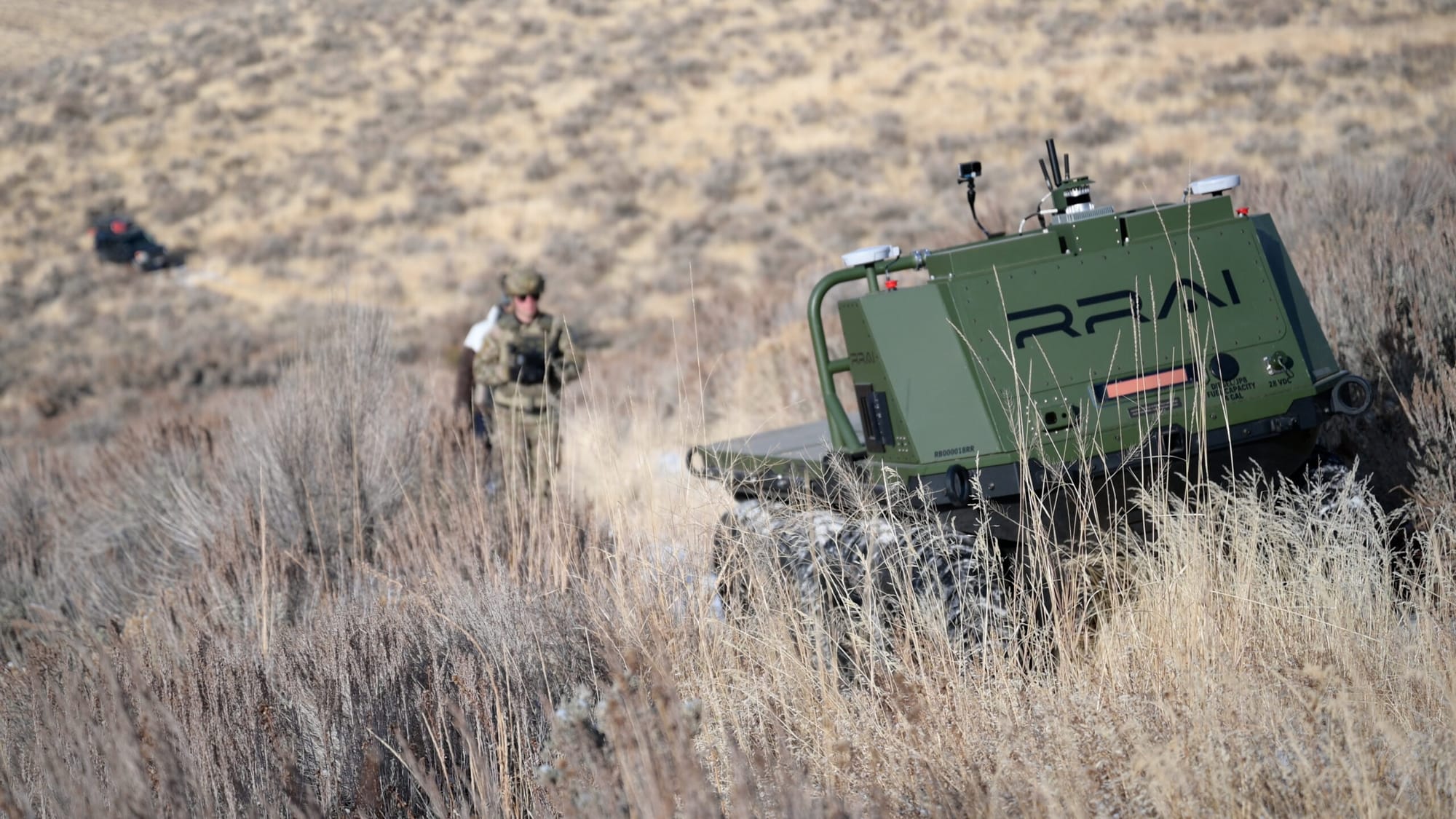 An olive green unmanned military ground vehicle with RRAI branding traversing dry grassy hills while soldiers follow behind.