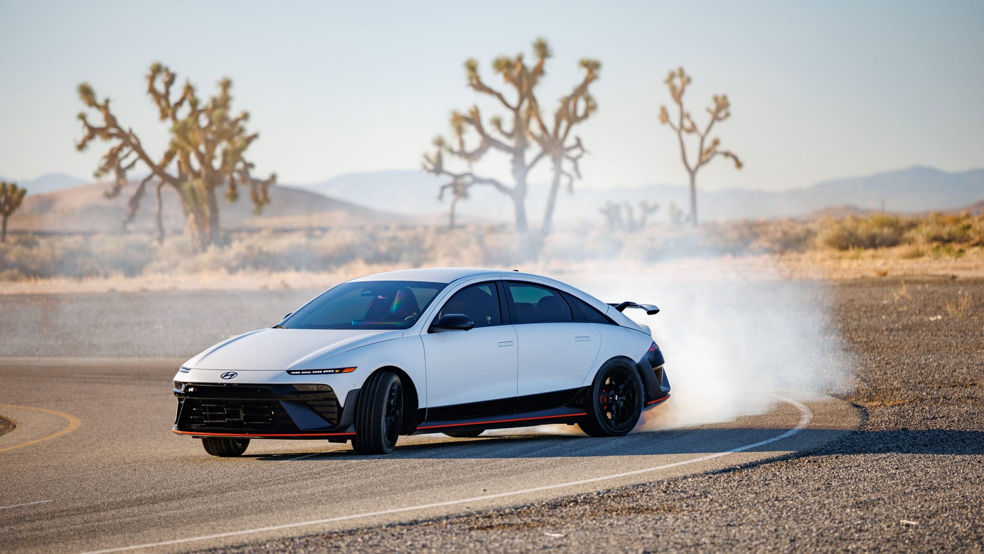 A white Hyundai performance electric sedan drifting on an asphalt track in a desert landscape with smoke billowing from its tires and Joshua trees in the background.